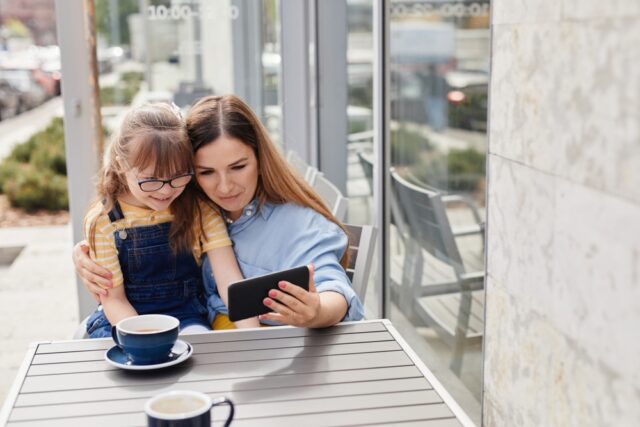 loving mother embracing girl with down syndrome while sitting at table in outdoor cafe together and looking at smartphone screen