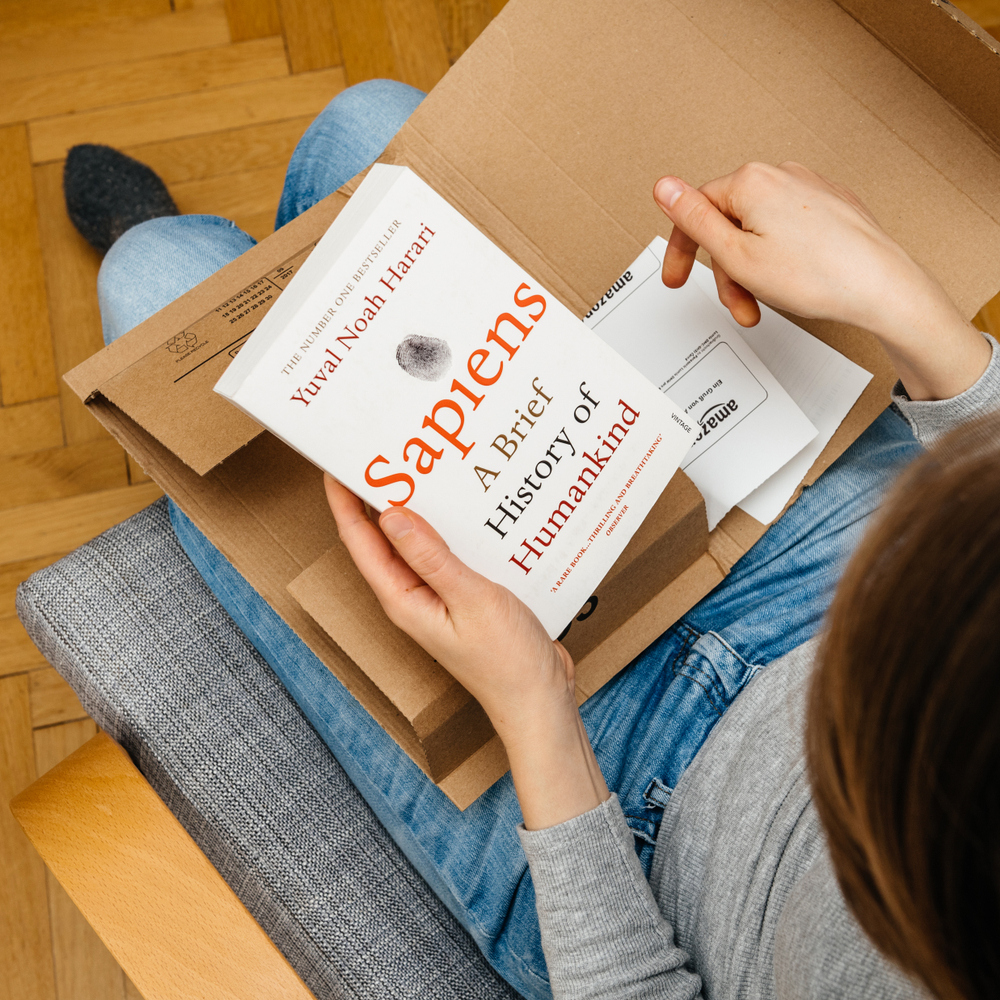 A woman pulling out Sapiens by Yuval Noah Harari from a box