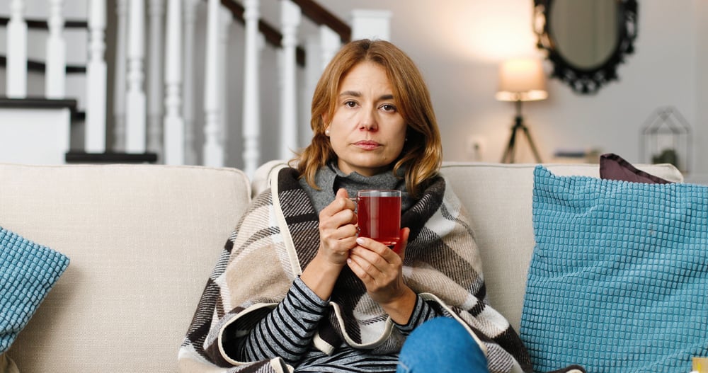 A sick matured woman sitting on the couch wrapped in a blanket and drinking a cup of hot tea