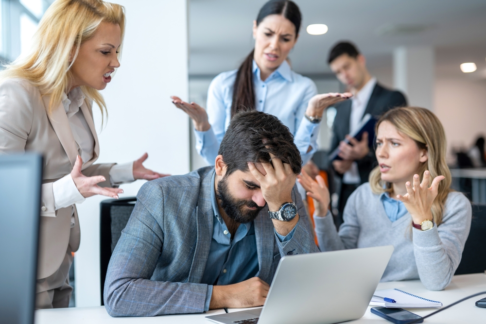 A frustrated businessman covering face with hands, surrounded by angry colleagues that are mad at him