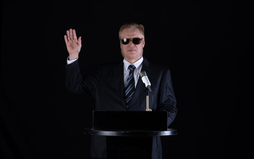 A male politician with microphone on podium and his right hand raised
