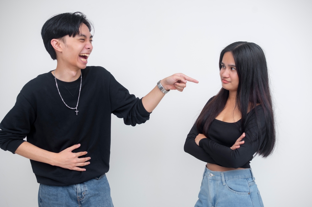 A young Asian man laughing and pointing at his girlfriend, making fun of her