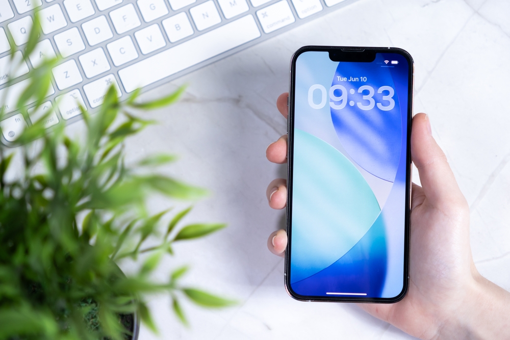 A man holding his iPhone close to his keyboard and his phone is showing the time and date on its locked screen