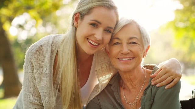 A young woman and an older woman smiling and embracing each other outdoors, with sunlight in the background and trees softly blurred—a heartwarming moment that beautifully captures the enduring bond between adult children and their parents.