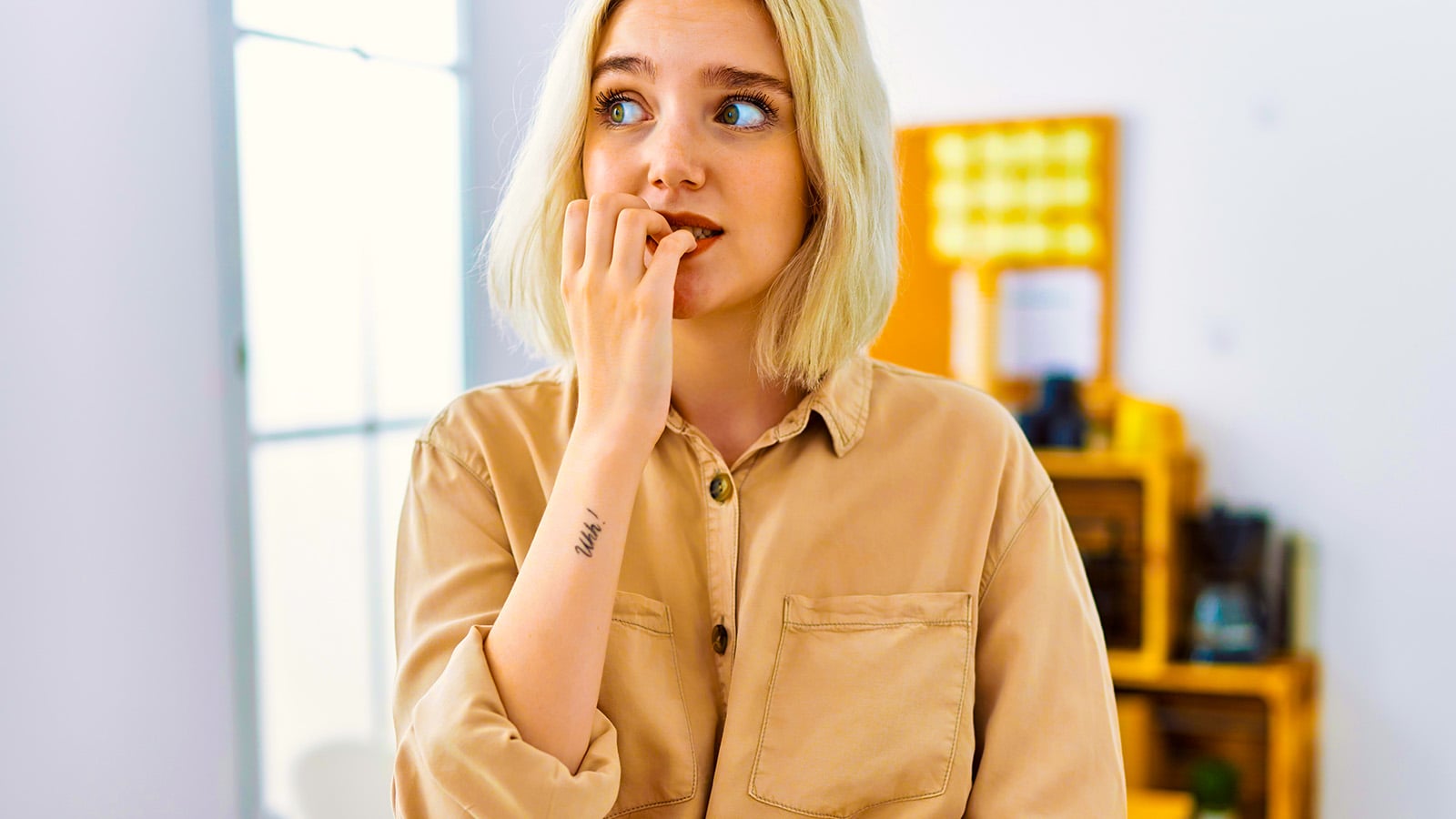 A person with blonde hair wearing a beige shirt stands indoors, nervously biting their fingernails and looking slightly to the side—an anxious gesture that psychology often links to adult patterns shaped by a lack of affection in childhood. The background is softly blurred with light and yellow decor.