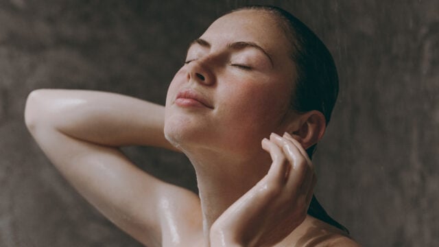 A person with closed eyes enjoys a relaxing night shower, tilting their head back with hands near their ears. Water droplets are visible on their skin and hair. The background is blurred and gray.