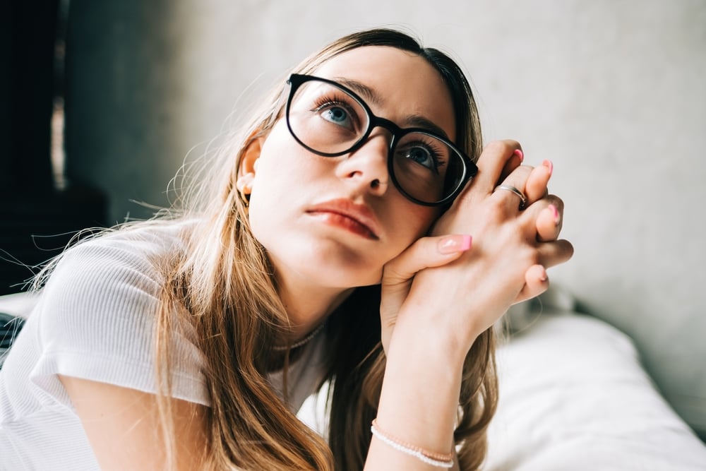 A young woman with long blonde hair and black-rimmed glasses looks upward thoughtfully, reflecting on independence as she rests her chin on clasped hands. She wears a white top and sits indoors with a soft, blurred background.