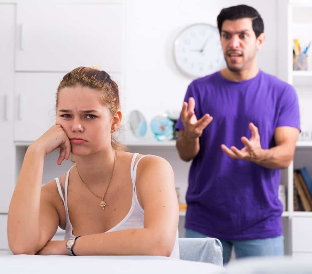 A frustrated girl sitting at home while her angry boyfriend yells at her and tries to dismiss her feelings