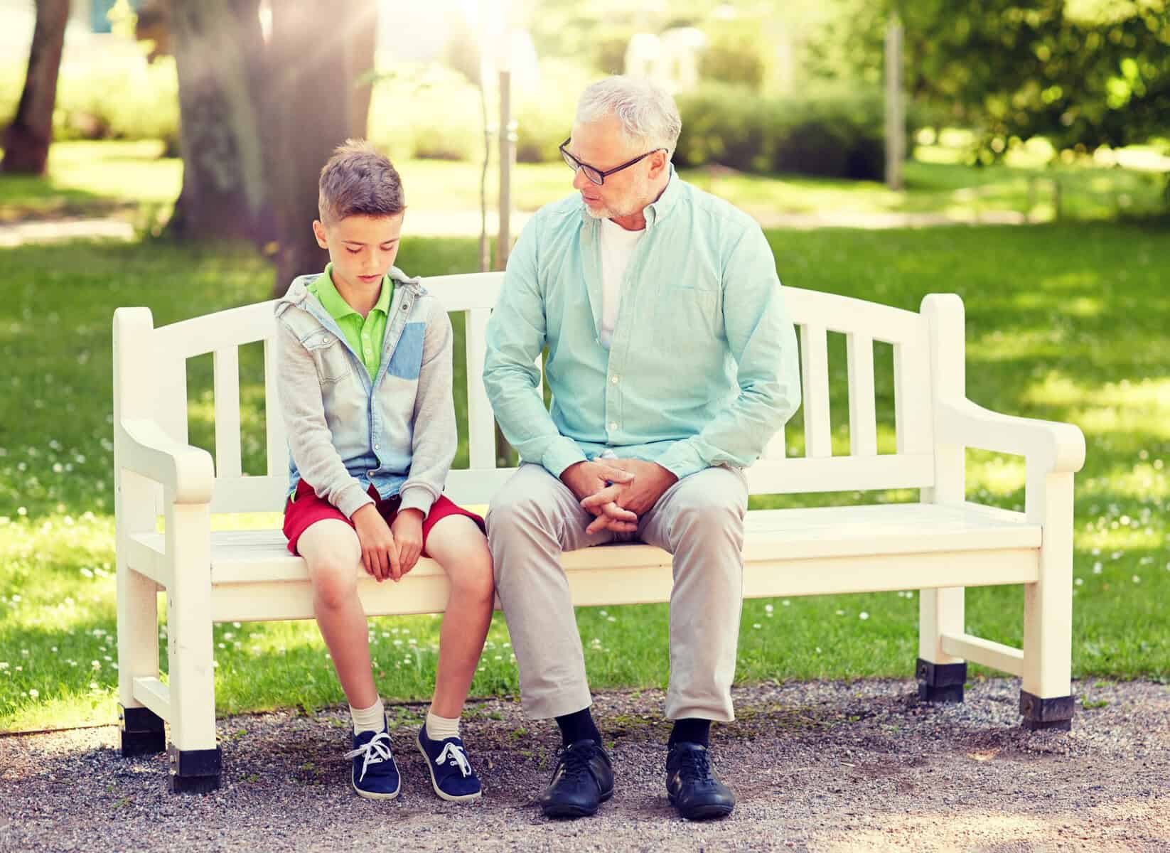 An unhappy little boy sitting on a park bench with his grandfather.