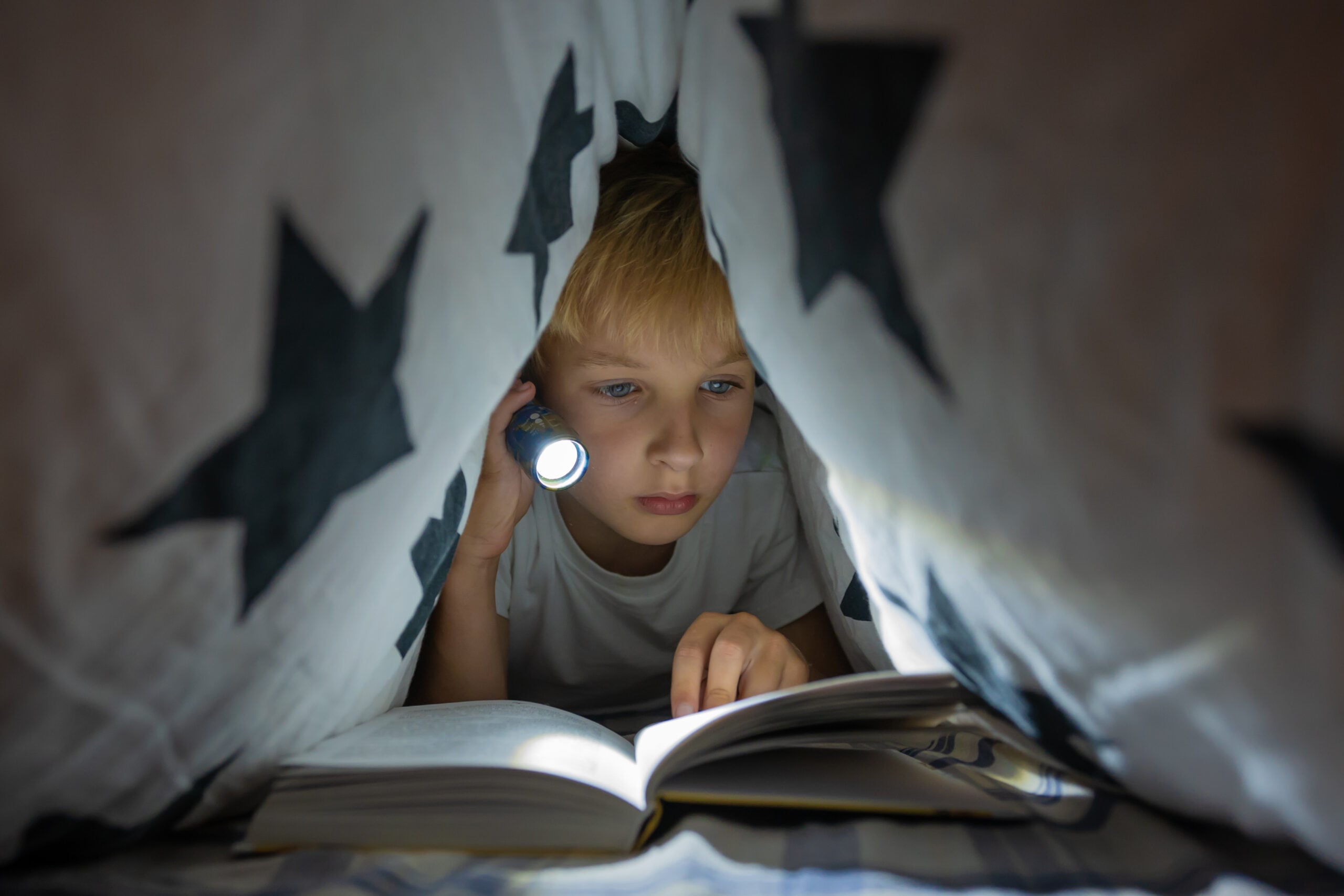 A little boy reading with a flashlight in a tent he made of blankets.