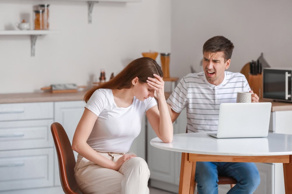 A young angry man working with his laptop is yelling and shouting at his girlfriend as they both sit at the kitchen table, the girlfriend is tired of the relationship