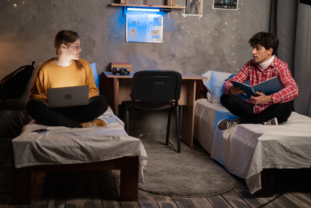 A male and female students sitting on their separate beds with one reading a book and the other with his laptop