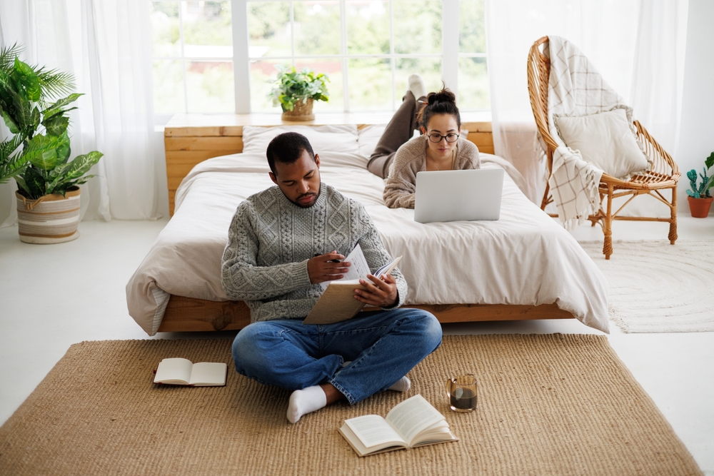 A young couple together in a room, the man is seated on the ground and reading and the woman is lying on the bed and working with her laptop