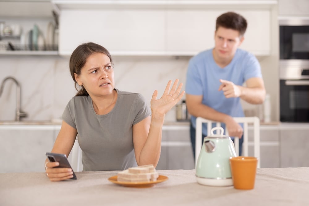 An offended upset girl having a quarrel with a young man behind her in the kitchen