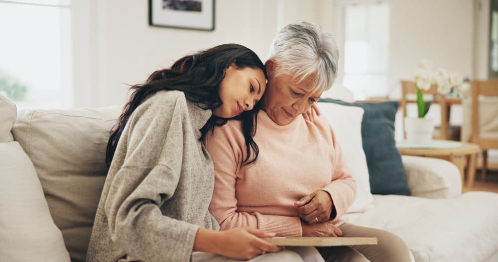 An adult daughter resting her head on her elderly mother's shoulder.