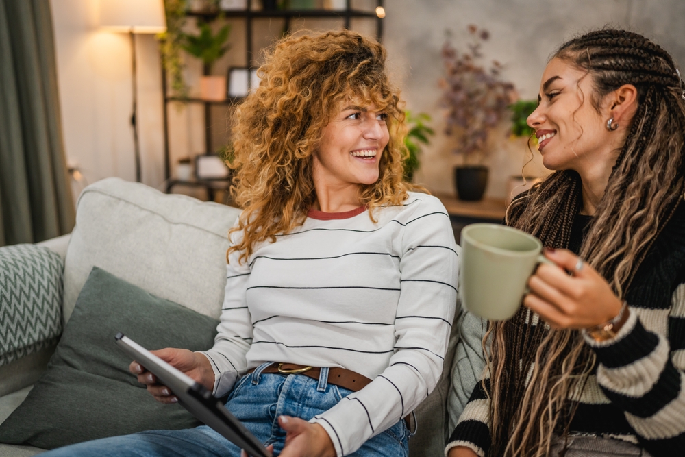 Two cheerful female friends talking and having coffee on the couch