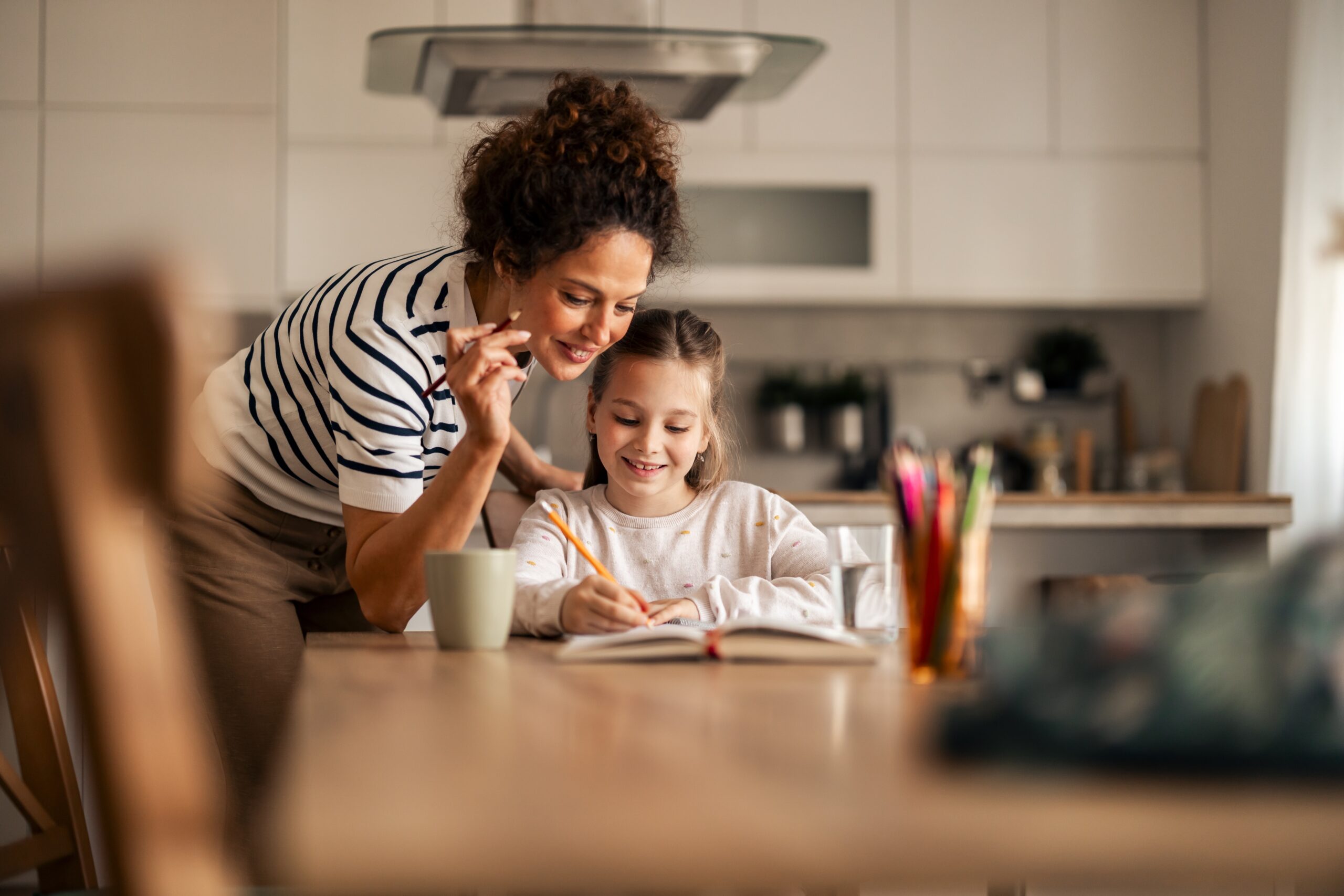 A mother happily helping her young daughter with schoolwork.