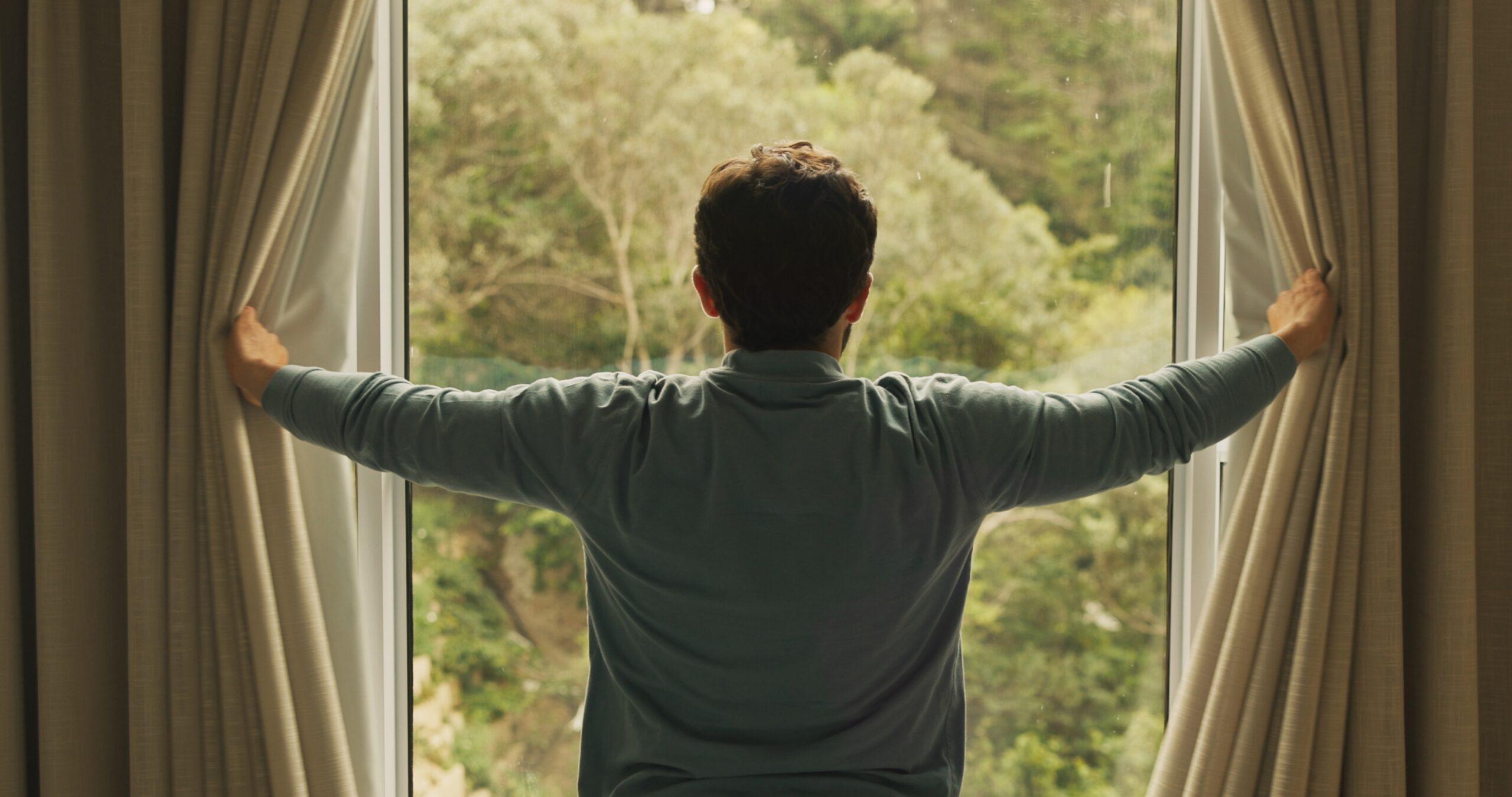 Man waking up and looking out of window towards a lush view.