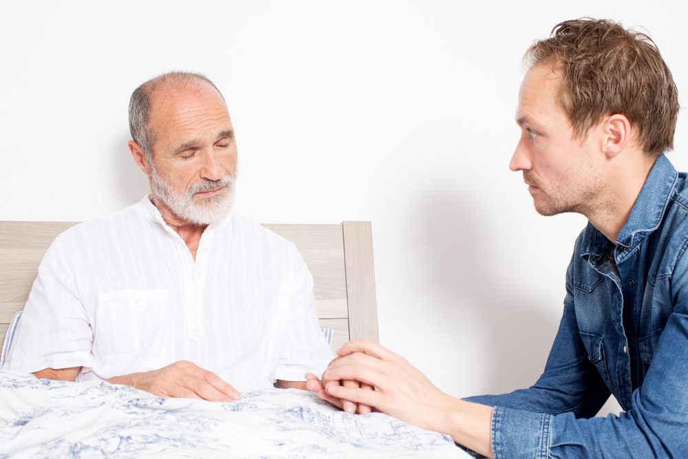 A senior man sitting up in bed and talking to his mature son