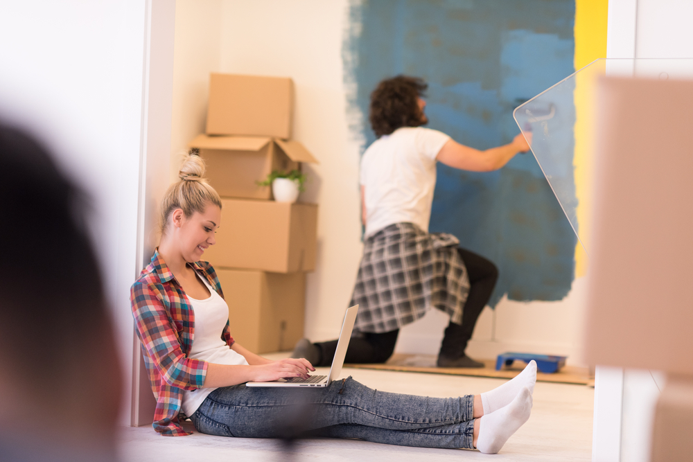 Two people together in a room, one is painting the wall and the other is seated on the floor with her laptop and working with it