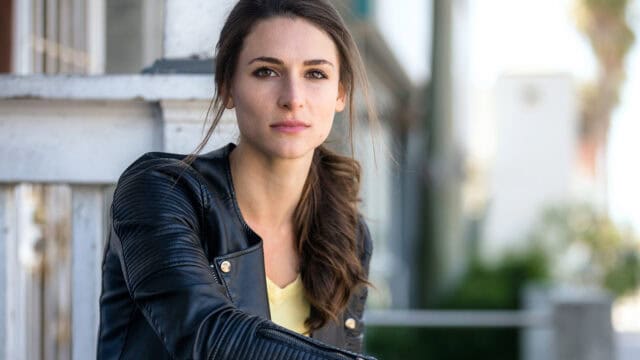 A woman with long brown hair in a ponytail, wearing a black leather jacket and yellow top, sits outdoors near a white wooden railing, looking calmly at the camera—her poised demeanor reflecting the quiet confidence often observed by psychologists in women who have moved beyond their people pleaser tendencies.