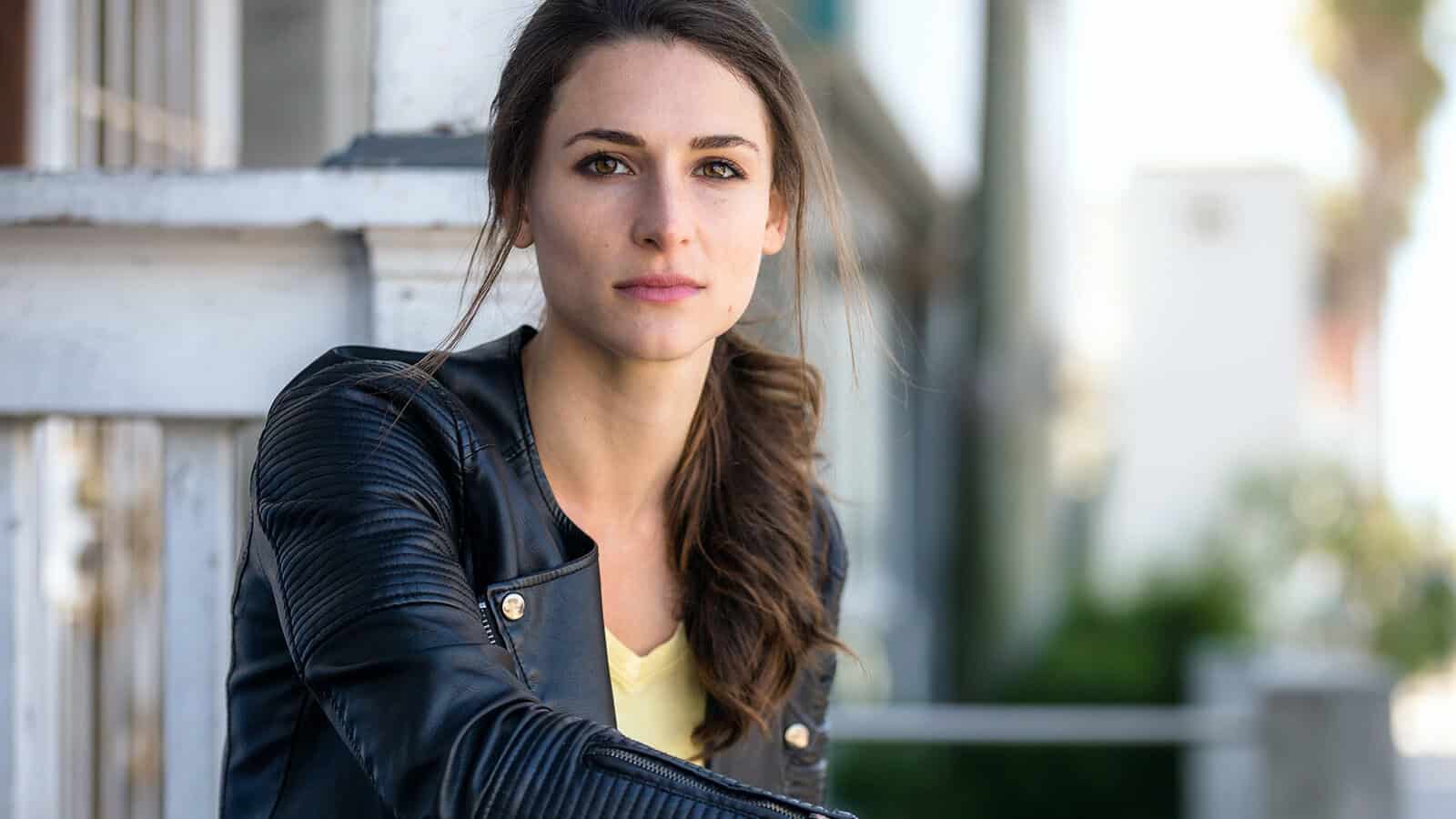 A woman with long brown hair in a ponytail, wearing a black leather jacket and yellow top, sits outdoors near a white wooden railing, looking calmly at the camera—her poised demeanor reflecting the quiet confidence often observed by psychologists in women who have moved beyond their people pleaser tendencies.