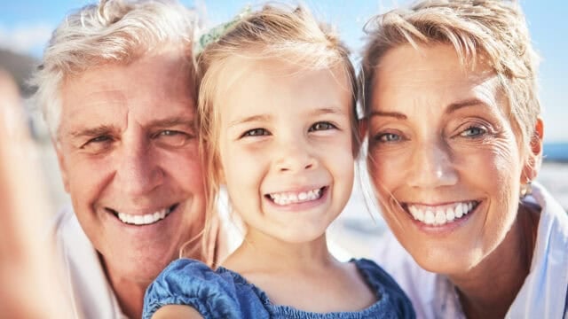 Smiling grandparents and a young girl pose closely outdoors, all looking at the camera. The group appears happy and cheerful, suggesting a special family moment together, possibly during a family trip to the beach or on vacation.