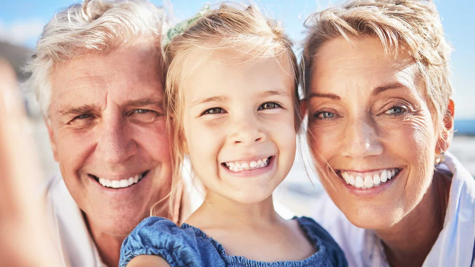 Smiling grandparents and a young girl pose closely outdoors, all looking at the camera. The group appears happy and cheerful, suggesting a special family moment together, possibly during a family trip to the beach or on vacation.