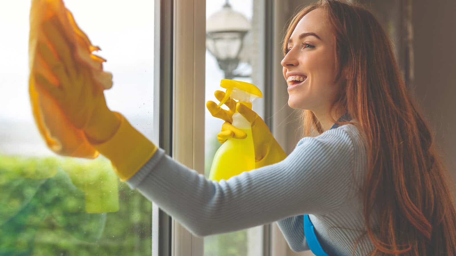 A woman wearing yellow rubber gloves smiles as she cleans a window with a yellow cloth and spray bottle, demonstrating her disciplined cleaning habits. Greenery and outdoor light are visible through the glass.