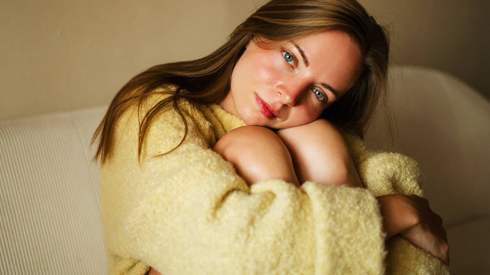 A woman with long brown hair and blue eyes sits on a couch, hugging her knees and wearing a cozy yellow sweater, looking at the camera with a gentle smile that hints at both vulnerability and quiet strength.