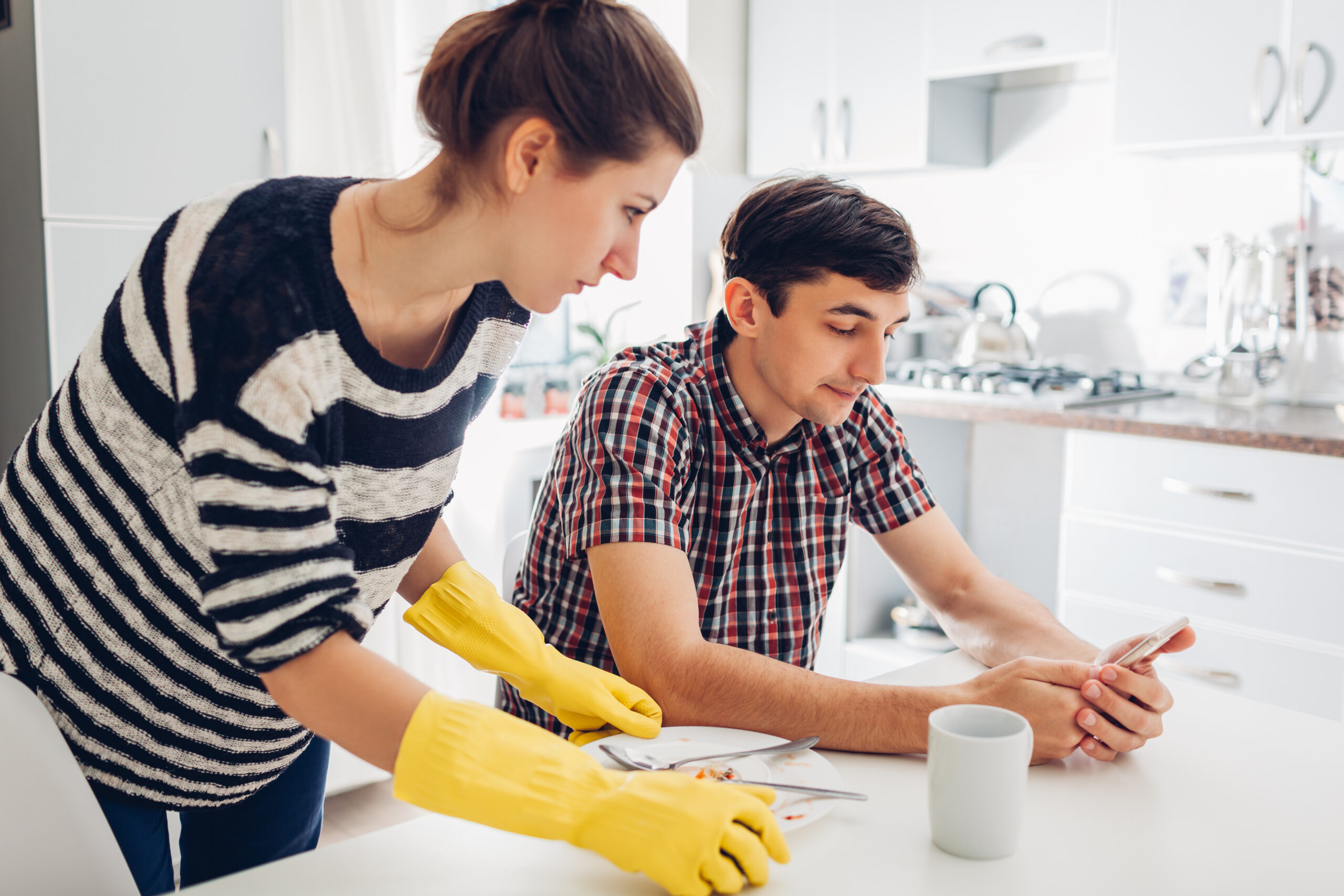 Lazy boyfriend on his phone while his girlfriend cleans his dishes.