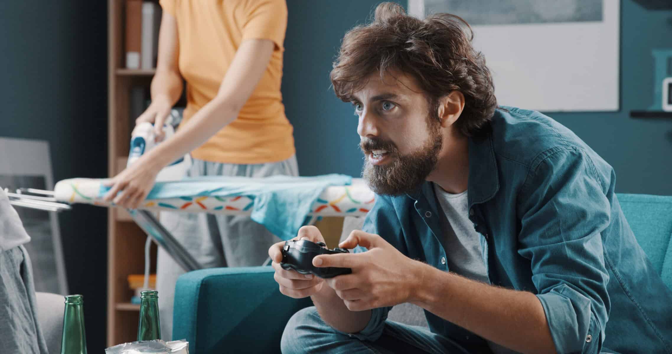 Lazy husband playing video games on the sofa while his wife is doing chores.
