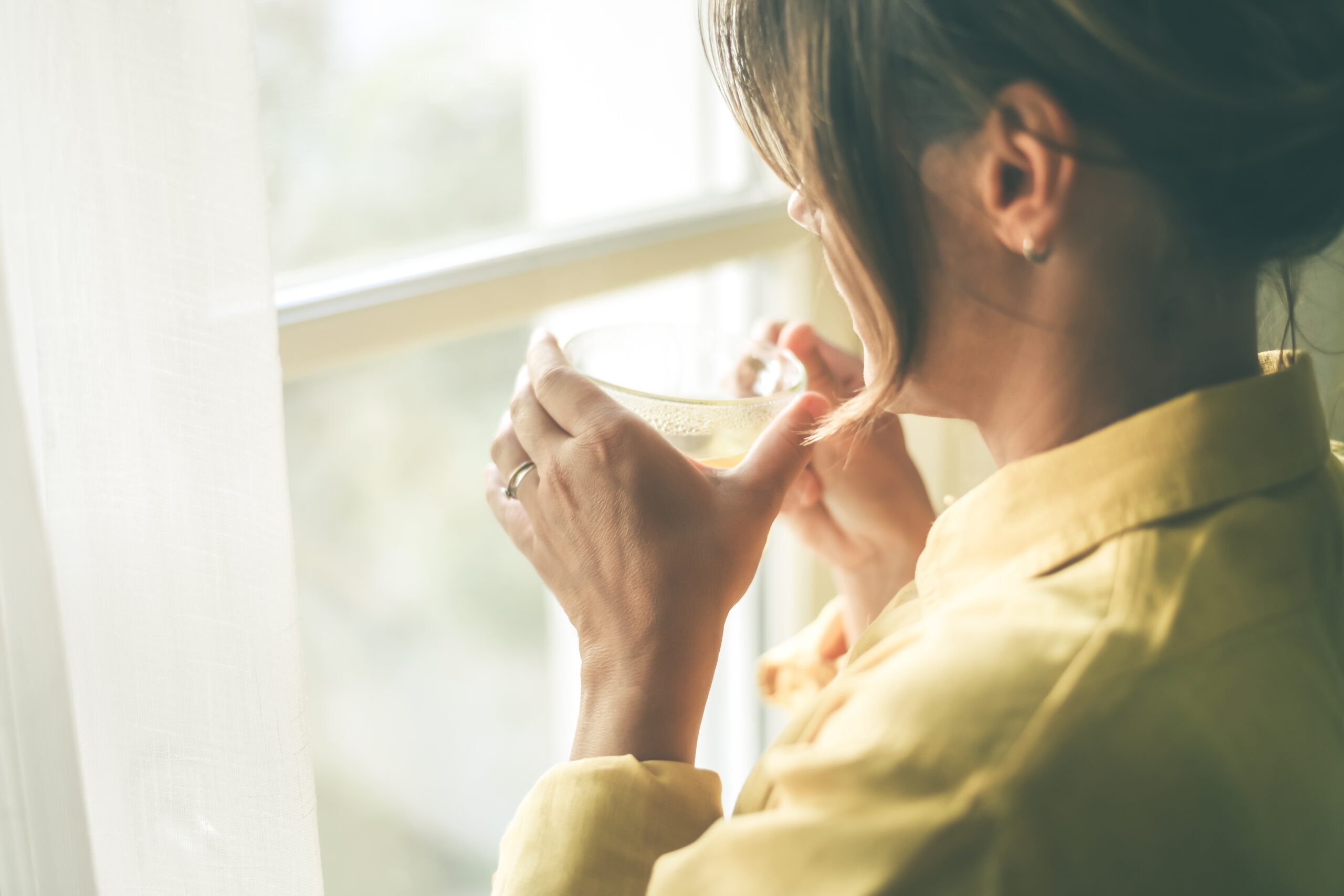 A woman alone drinking a cup of tea looking outside the window.