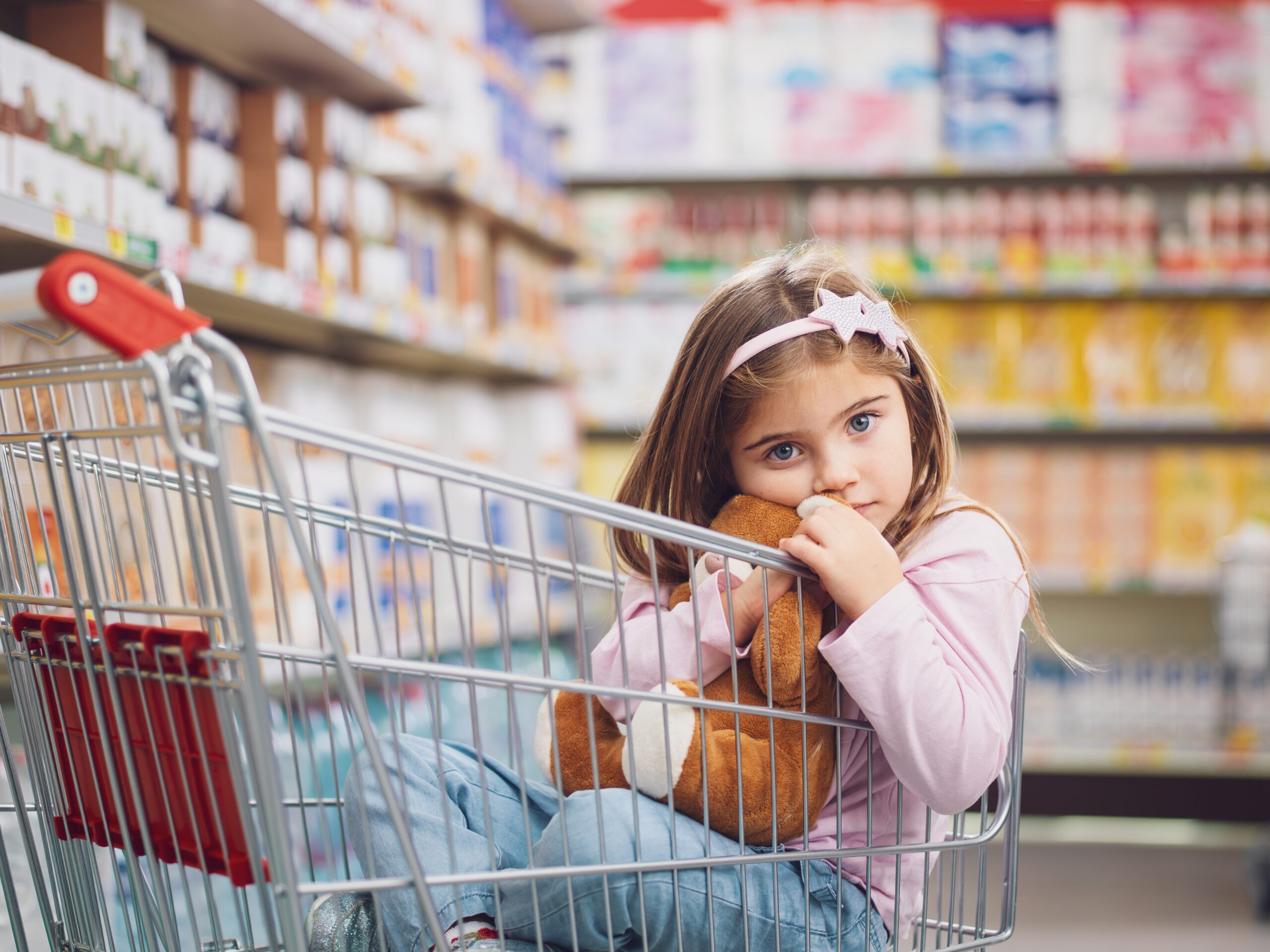 A little girl at the supermarket sitting inside of the cart while her mother shops.