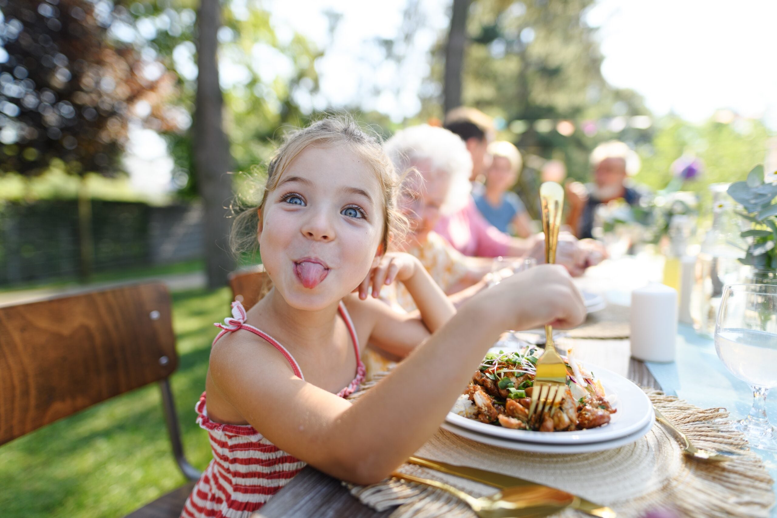 A funny little girl sitting at a table eating outdoors with her grandparents and parents.