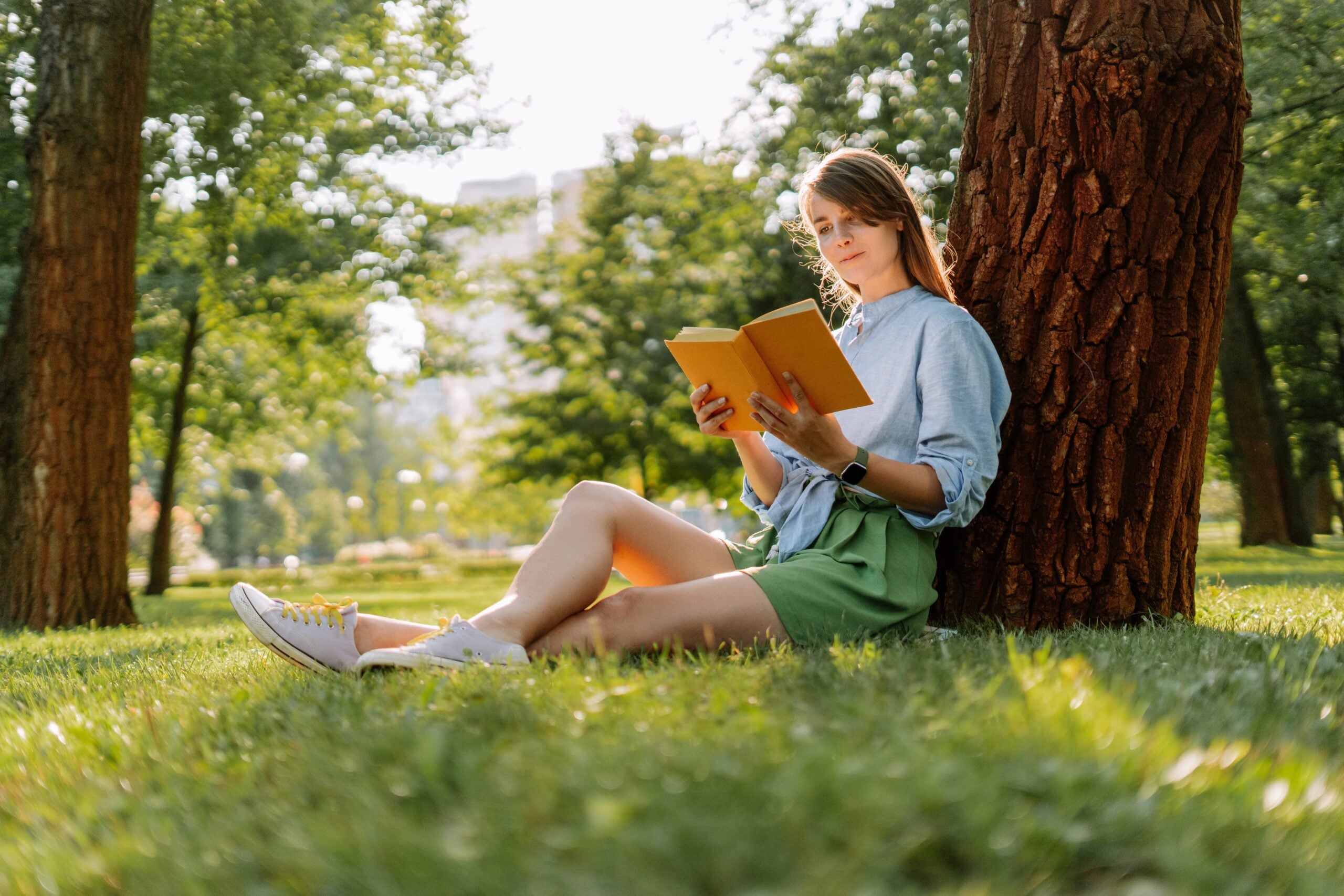 A woman reading peacefully in the park under a tree on a sunny day.