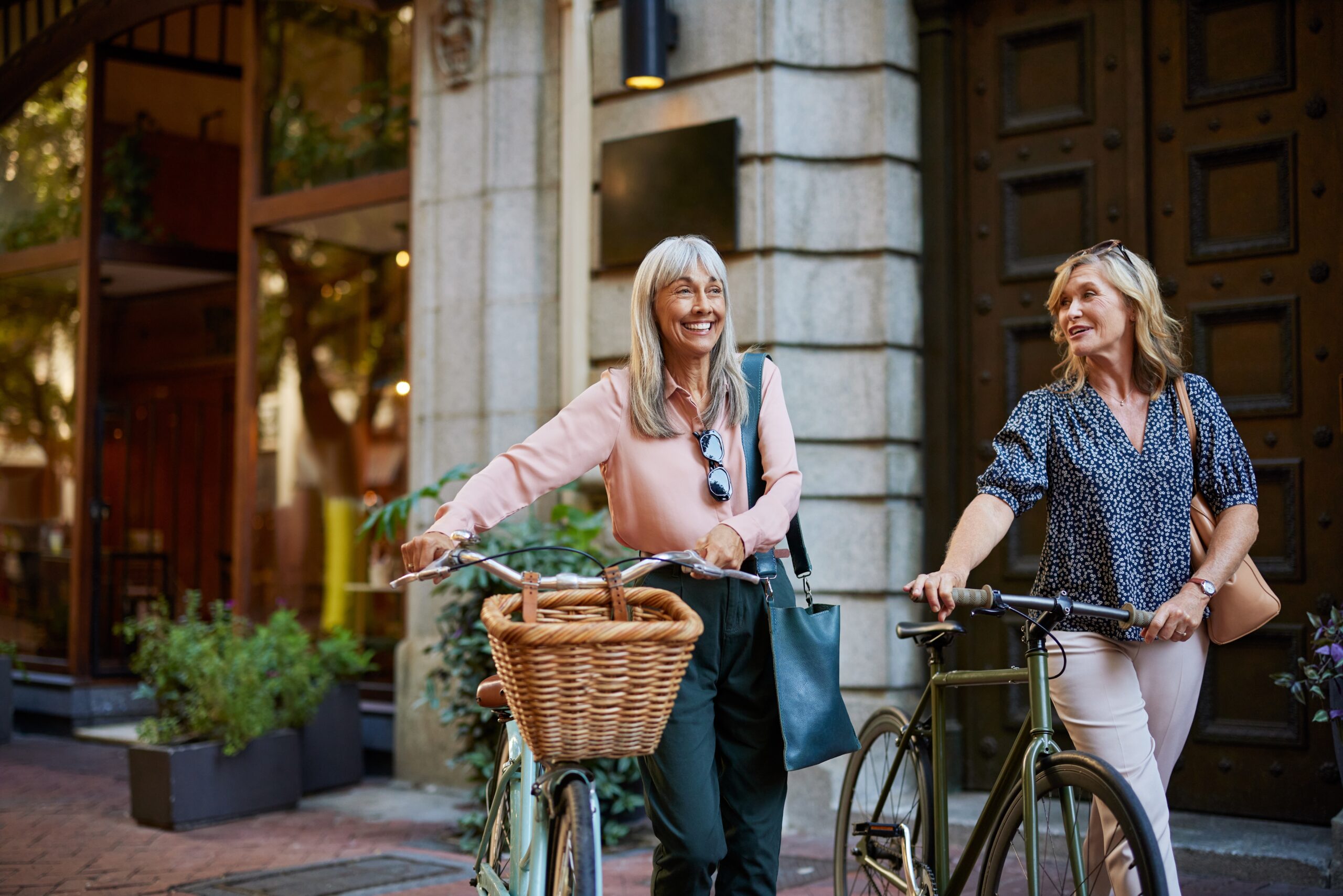 Two women smiling and pushing their bicycles outdoors.
