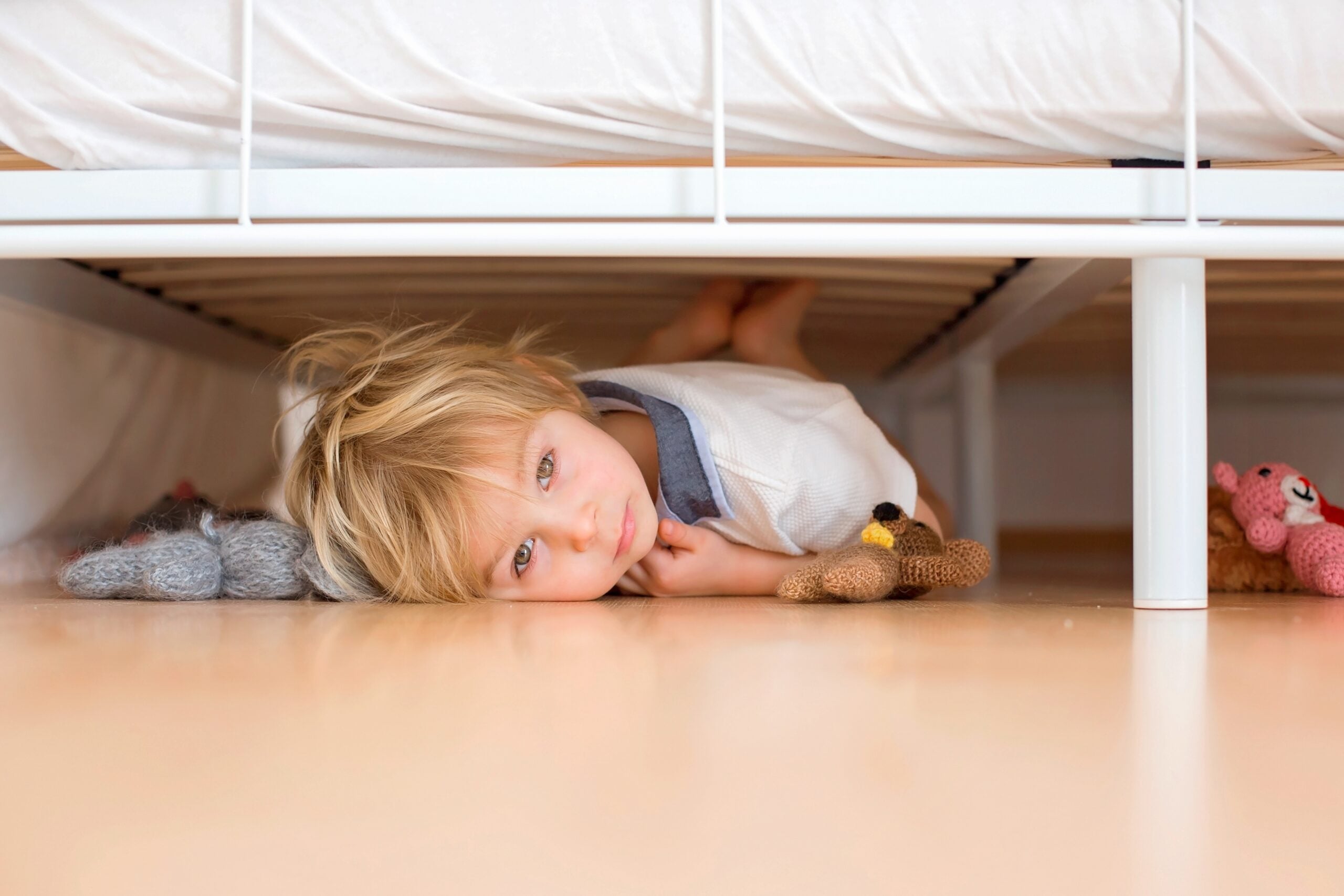 A frightened toddler hiding under the bed with his stuffed toys.