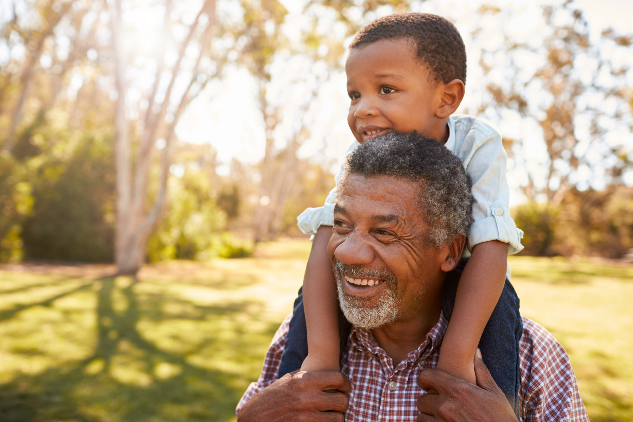 A smiling grandfather carrying his grandson on his shoulders in the park.