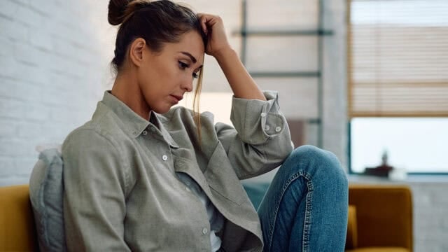A woman sits on a couch indoors, looking down with a thoughtful or worried expression, resting her head on her hand as if lost in overthinking.