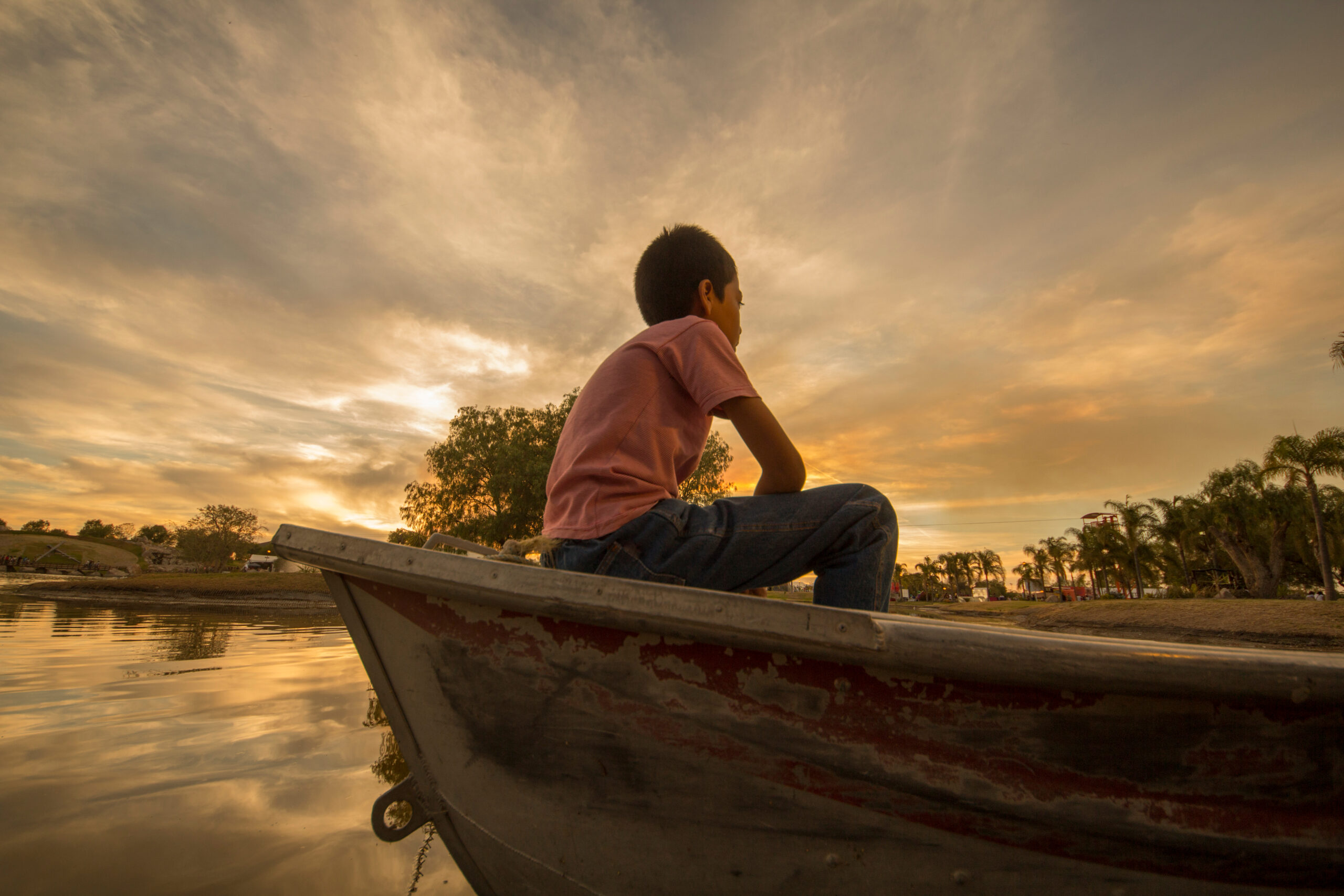 A boy in a boat by himself watching the sun set.