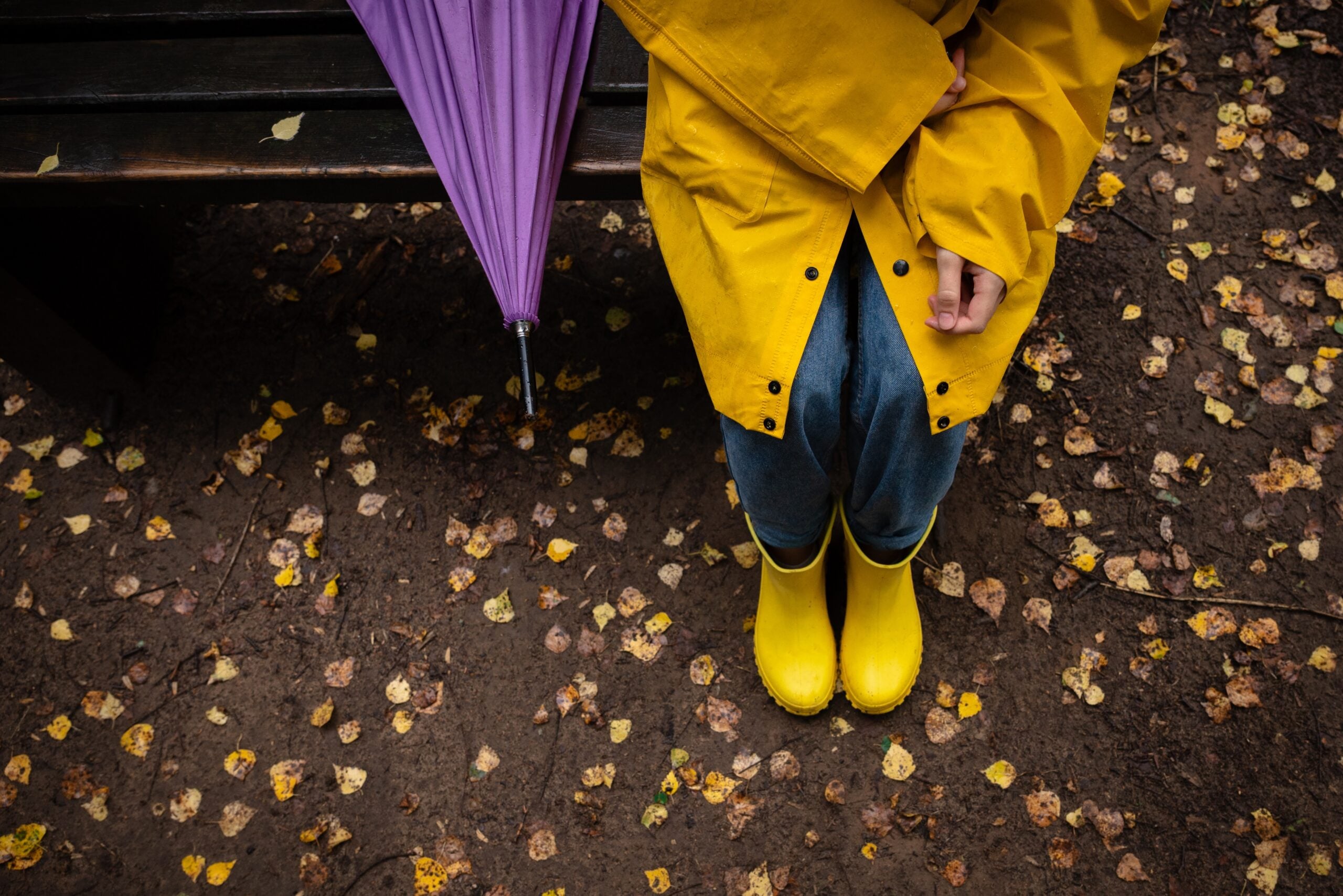 A woman in a yellow raincoat and rubber boots sits alone on a bench.
