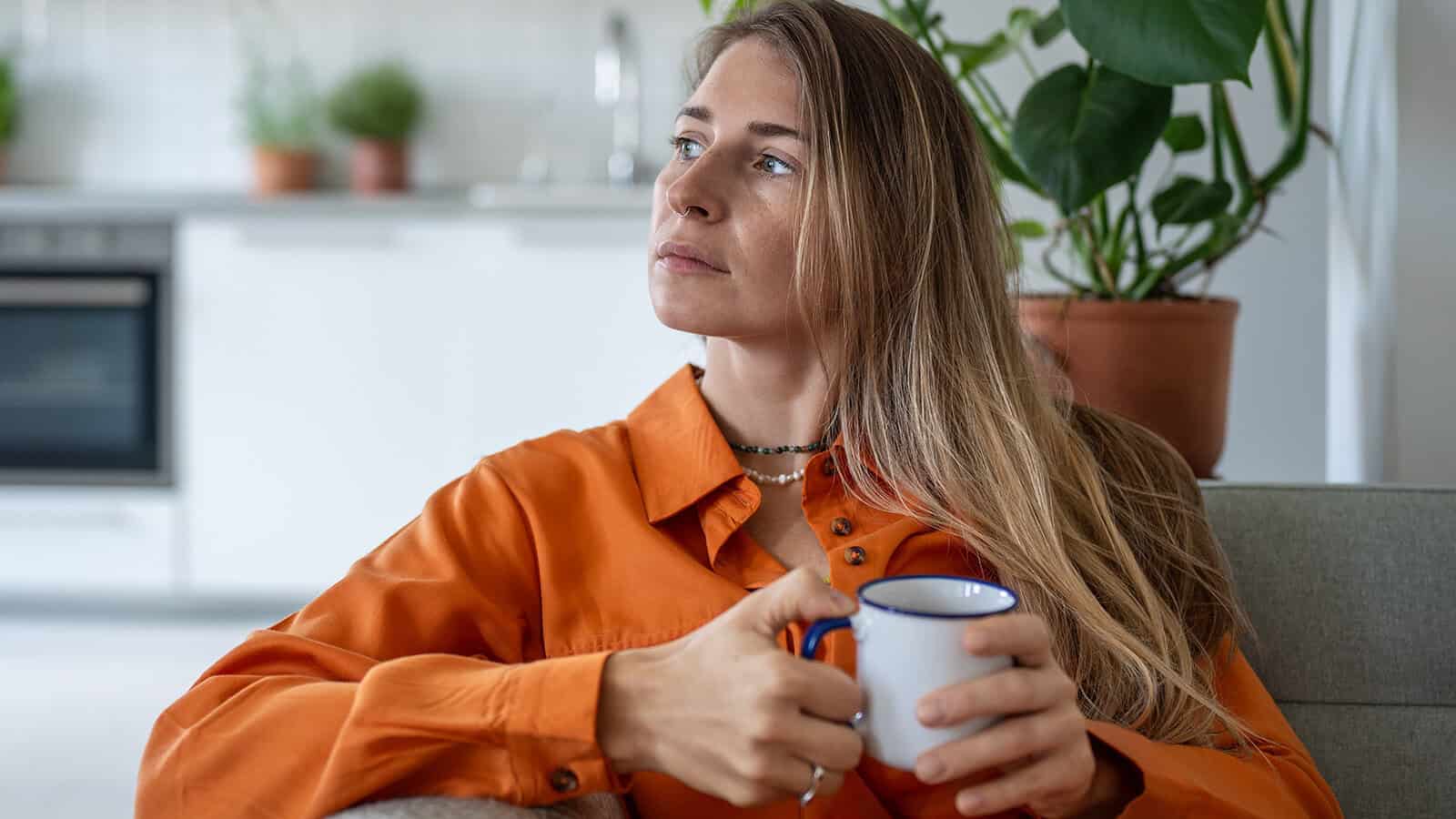 Thoughtful middle aged woman sits on sofa at home holding cup in hand looks towards window daydreaming. Peaceful pensive female thinking about future in cozy apartment in moment of quiet introspection