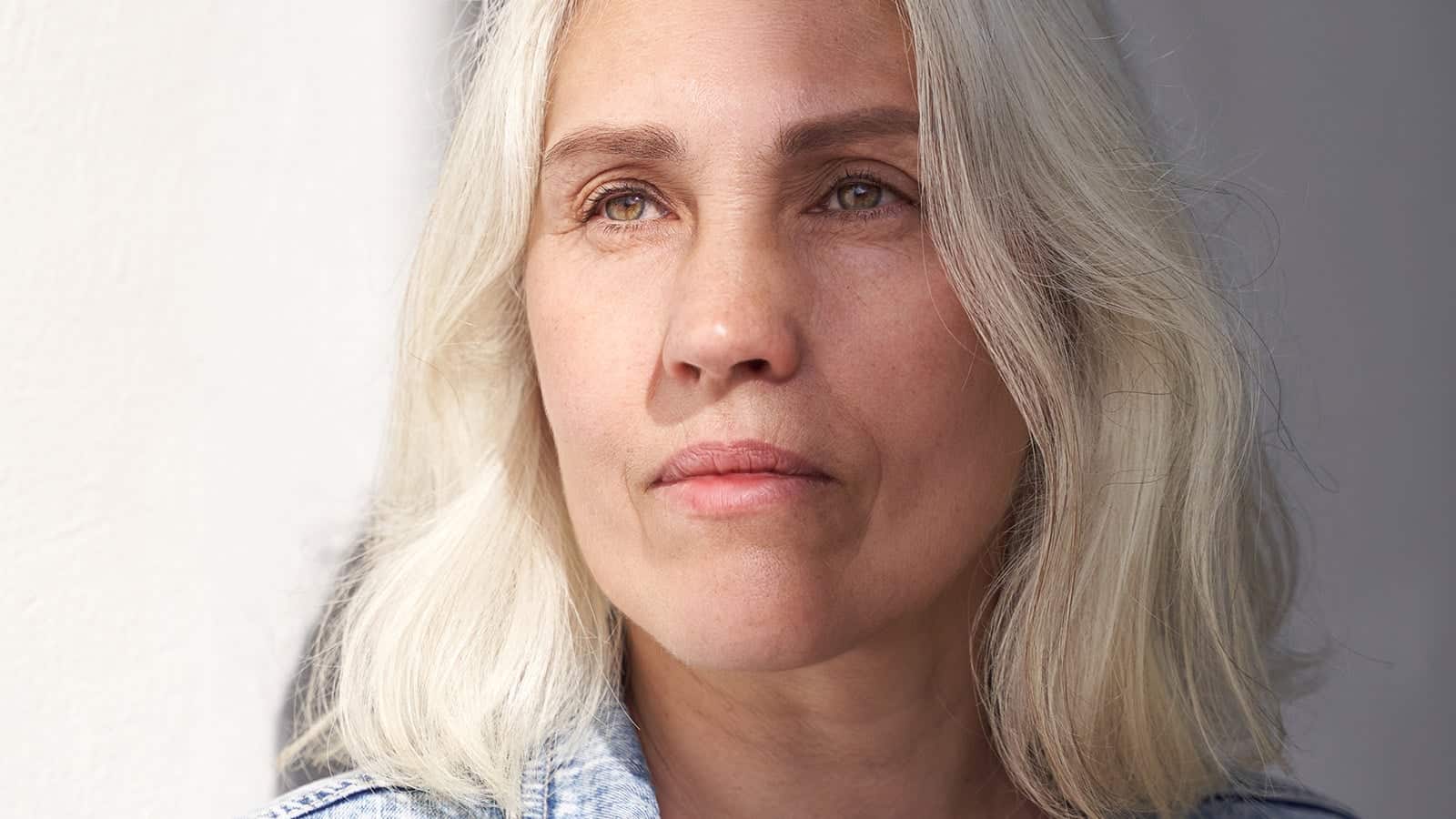 Mature woman with gray hair in relaxed denim outfit reflecting in natural light against minimalist background.
