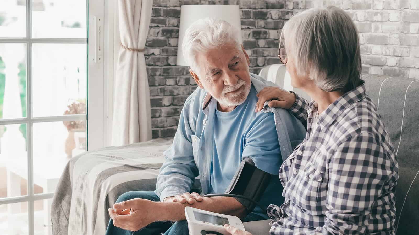Senior woman using medical device to measure blood pressure of her husband suffering of hypertension - elderly couple sitting at home on sofa takes care of health