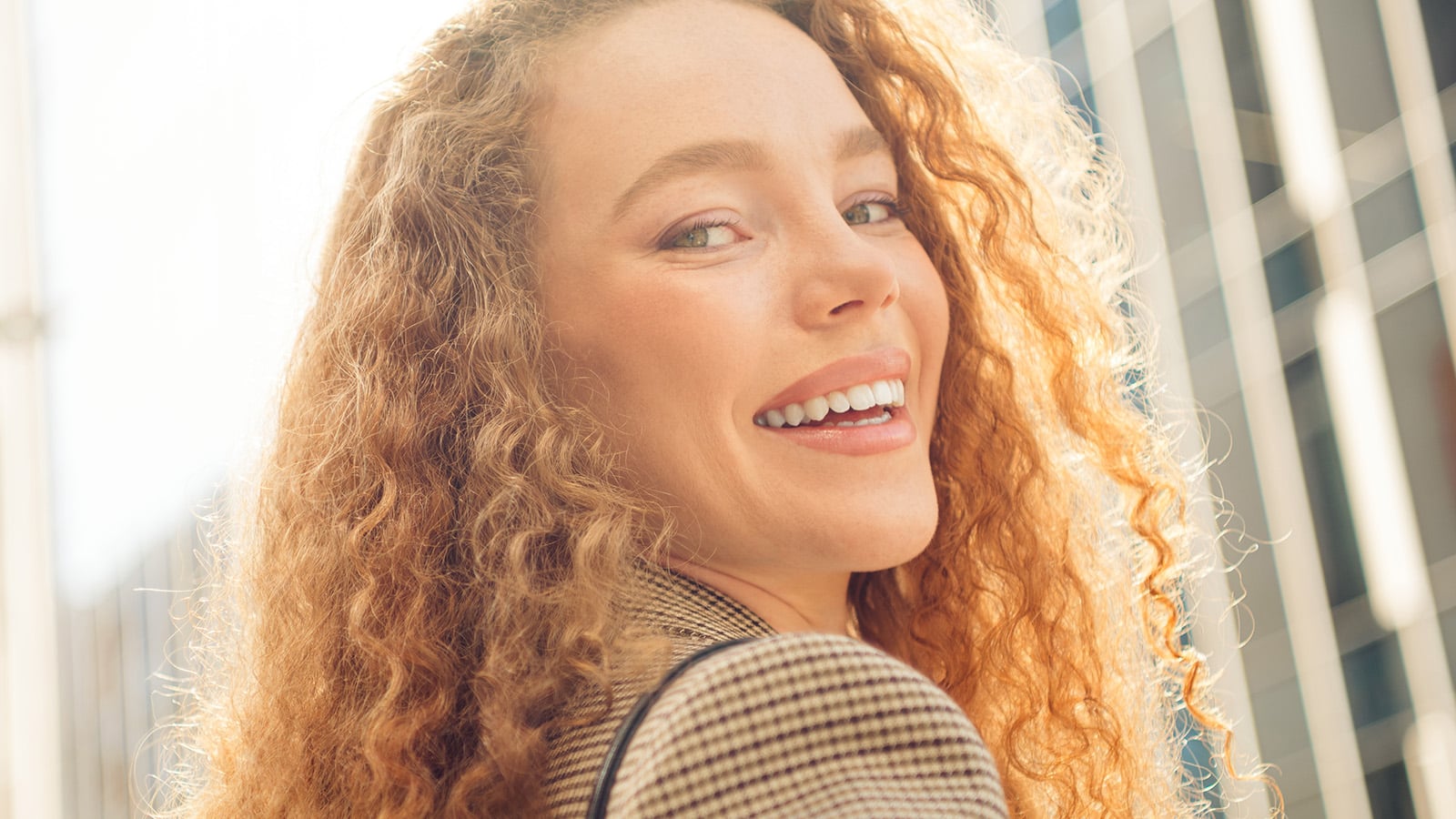 Young professional woman with red curly hair smiling outdoors in a modern cityscape, exuding confidence and happiness