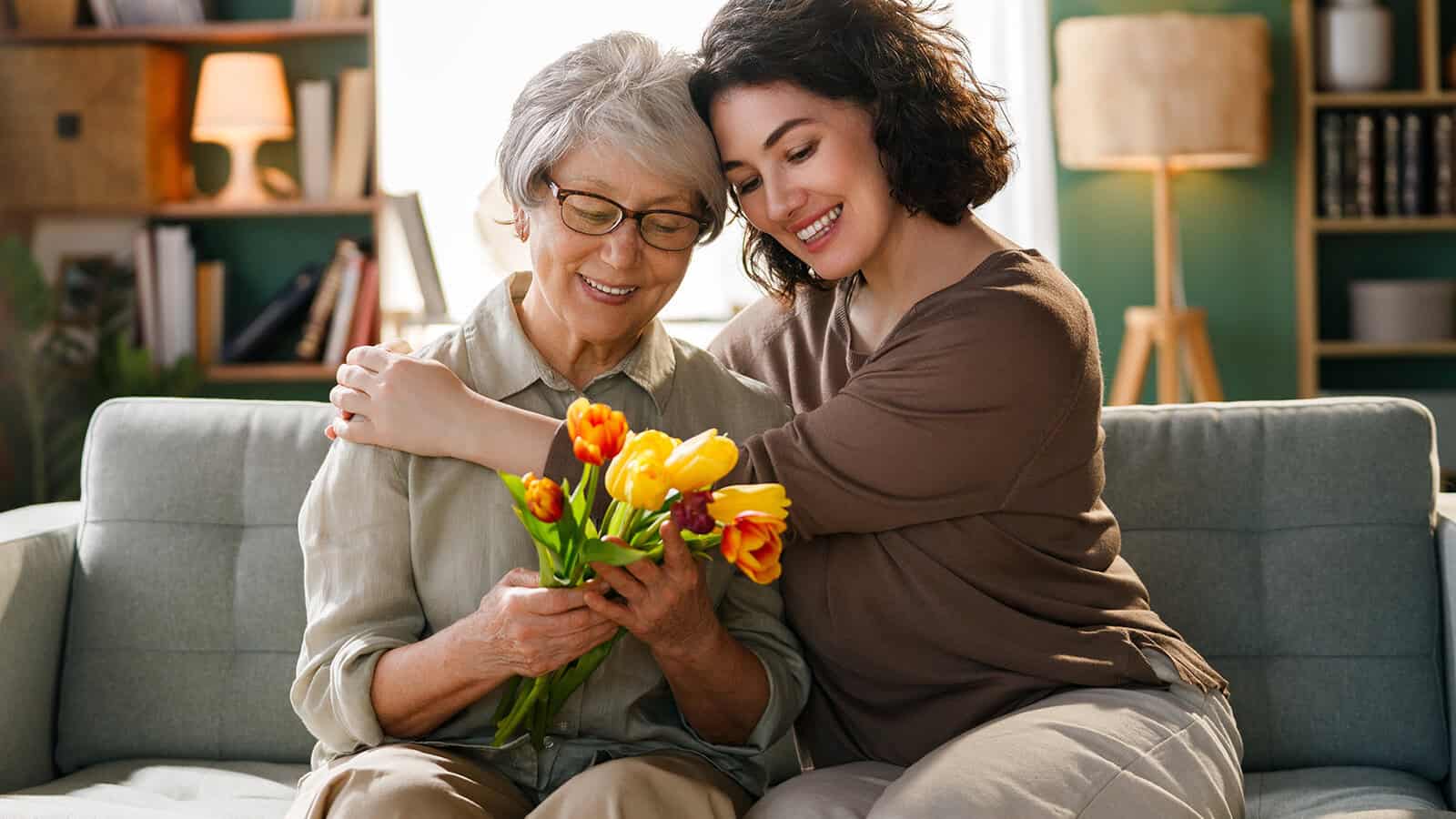 Beautiful tender moment between senior mother and adult daughter sitting on sofa in cozy home, sharing a colorful bouquet of tulips. Warm smiles, affectionate embrace, and heartfelt connection highlight authentic family love, intergenerational support, and gratitude. Perfect for Mother's Day, senior care, family celebration, or lifestyle concepts emphasizing togetherness, well-being, and emotional wellness.