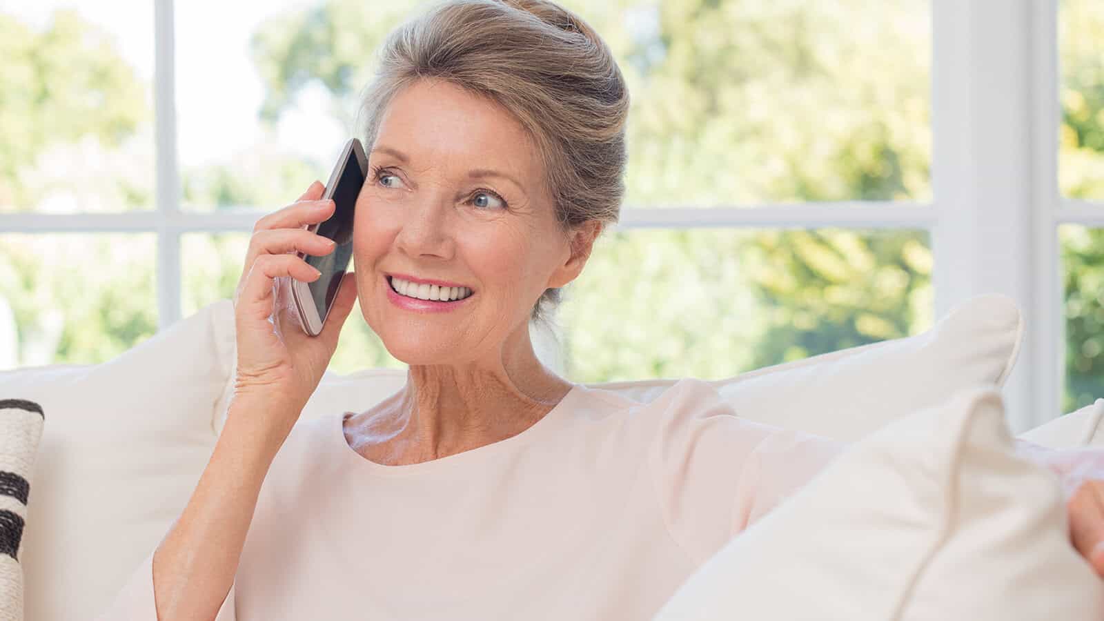 Senior woman talking on her mobile phone. Senior woman has a happy conversation at cellphone. Smiling senior woman using phone sitting on couch at home.