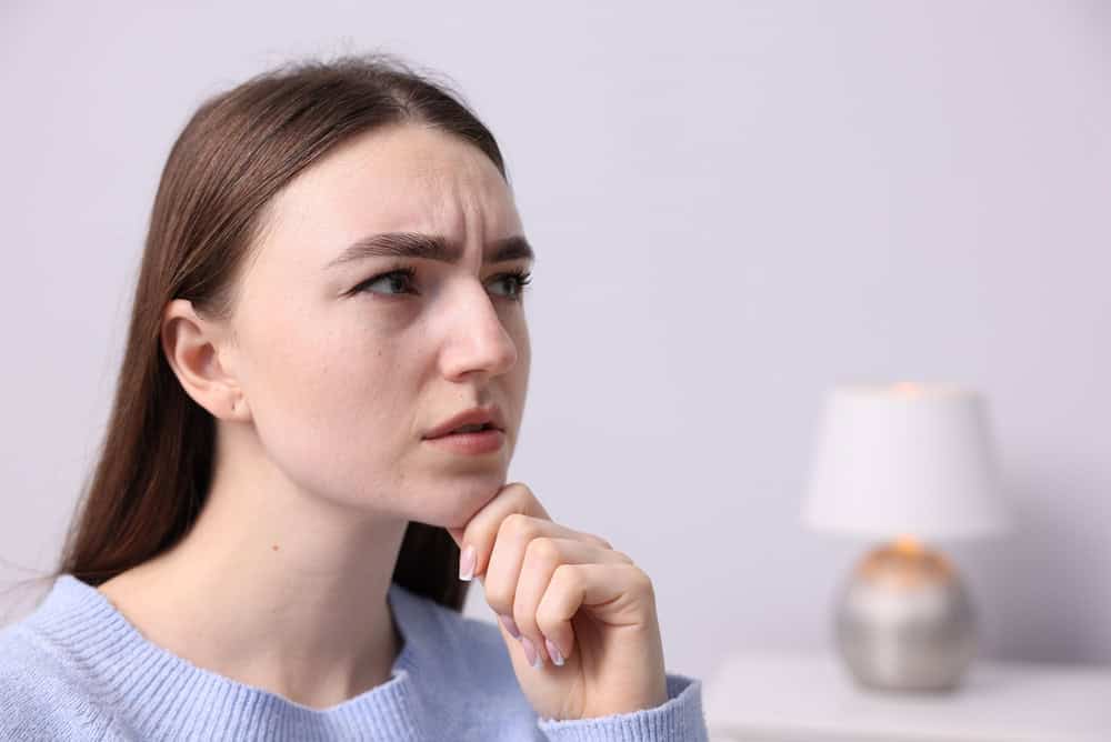 Young woman with long brown hair and a serious expression, resting her chin on her hand as if thinking deeply about psychology or remembering names, with a blurred lamp in the background.
