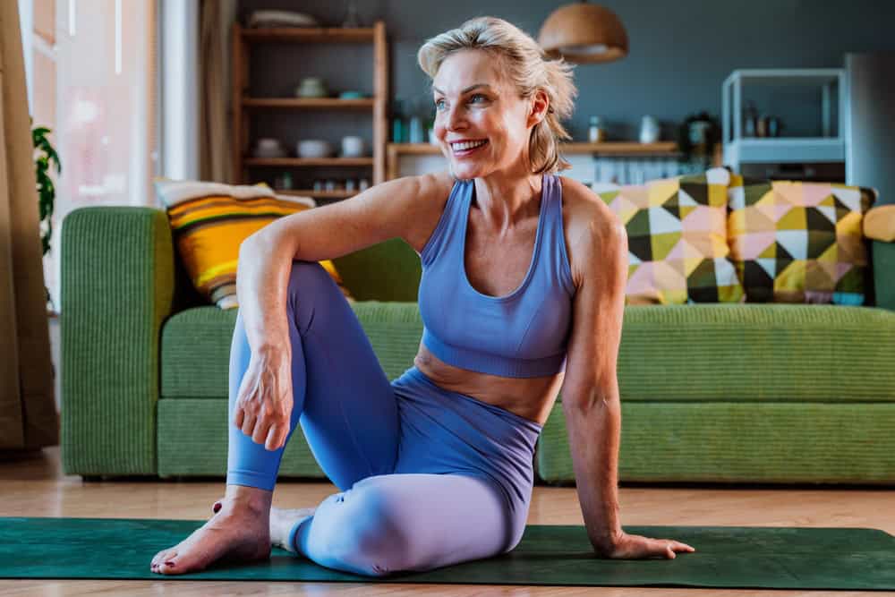 A smiling woman in a purple sports outfit sits on a yoga mat in her living room, radiating life satisfaction after exercising. A green couch with colorful pillows and shelves accent the cozy space, reflecting contented people in their 60s.
